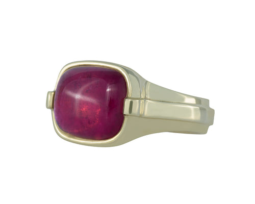 Large green gold ring with overlapping ribbon of gold around the band set with large rectangle cabochon of deep pink tourmaline. Gem lies across the finger.