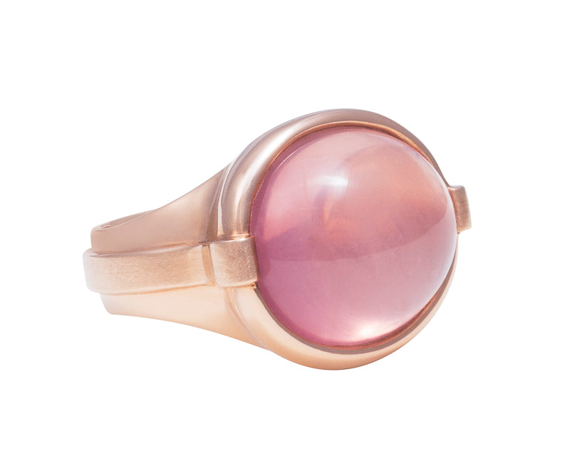 Large rose gold ring with overlapping ribbon of gold around the band set with large oval cabochon of rose quartz. Gem lies across the finger.