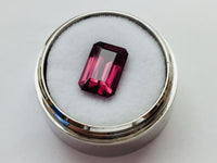 Medium size rectangular deep purple rhodolite garnet gem, on white background in gem jar.