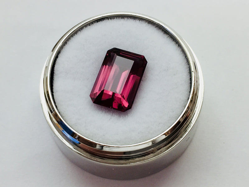 Medium size rectangular deep purple rhodolite garnet gem, on white background in gem jar.