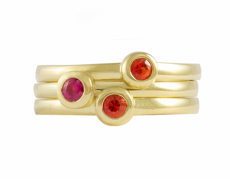 Three thin green gold rings stacked together. Each ring is set with a bright pink or orange sapphire that are set in a frame.
